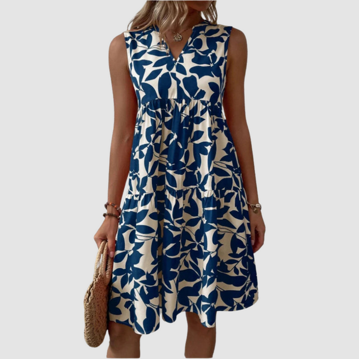 Model wearing a sleeveless blue and white floral print flowy dress, holding a woven bag, against a plain background.