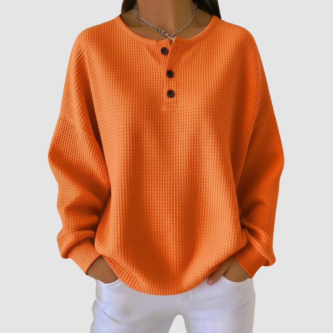 Orange relaxed sweater with buttoned neckline and textured fabric, worn with white pants in a studio setting.