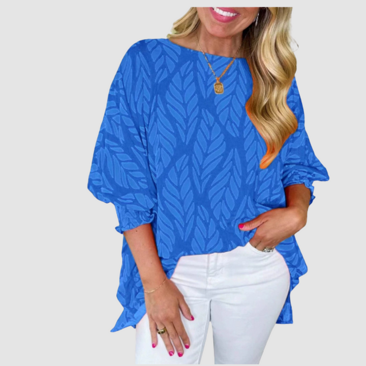 Woman wearing a blue loose blouse with leaf pattern and white pants in a studio setting.