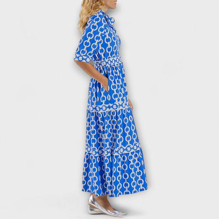 Side view of woman wearing blue and white chain print midi dress with hands in pockets, against a plain background.