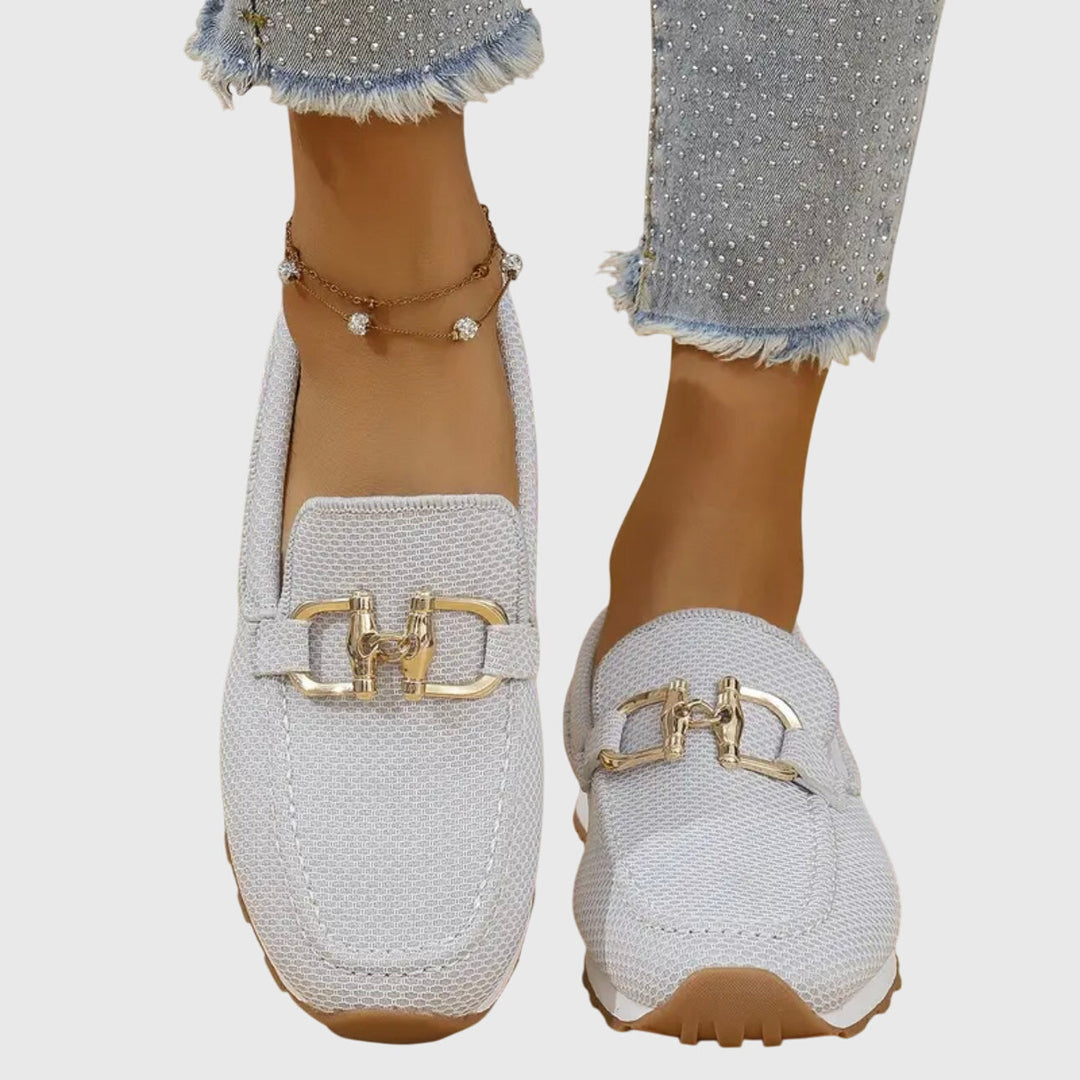 Light gray orthopedic loafers with gold detail worn with frayed jeans and anklet on a light background.