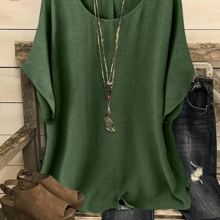 Olive green loose summer top on hanger with necklace, sandals, and jeans on rustic bench.
