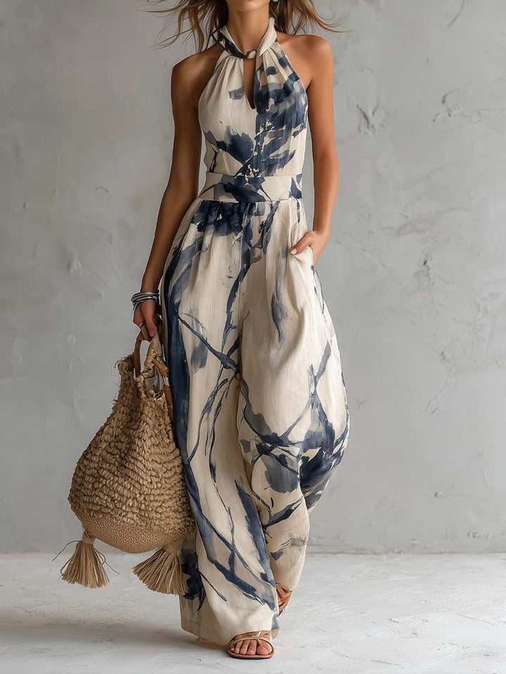 Flowy halter-neck jumpsuit with blue abstract print, beige background, boho bag. Studio setting with textured wall.