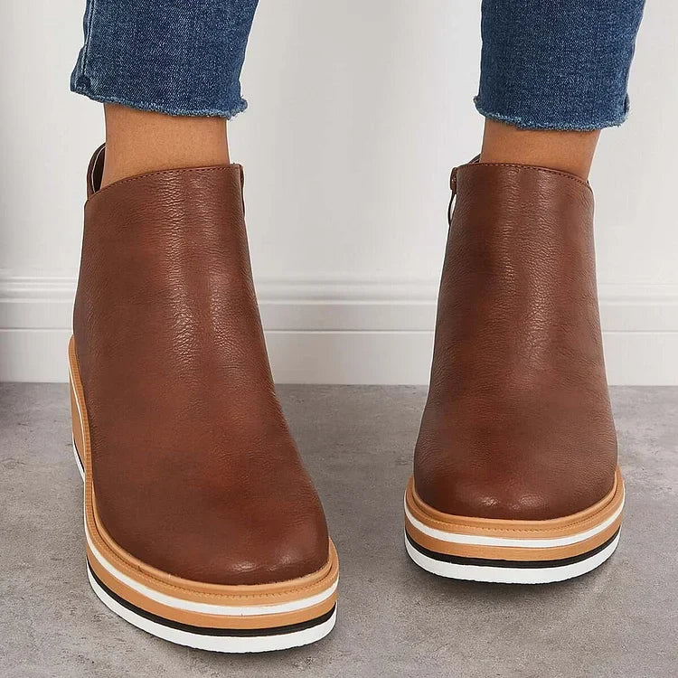 Front view of brown luxe boots with wedge soles, worn with frayed jeans, on a gray floor.