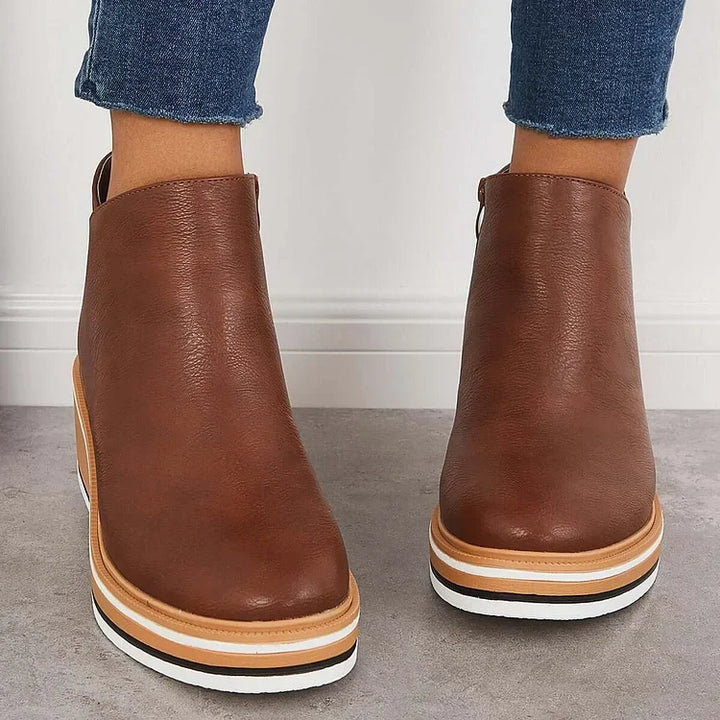 Front view of brown luxe boots with wedge soles, worn with frayed jeans, on a gray floor.