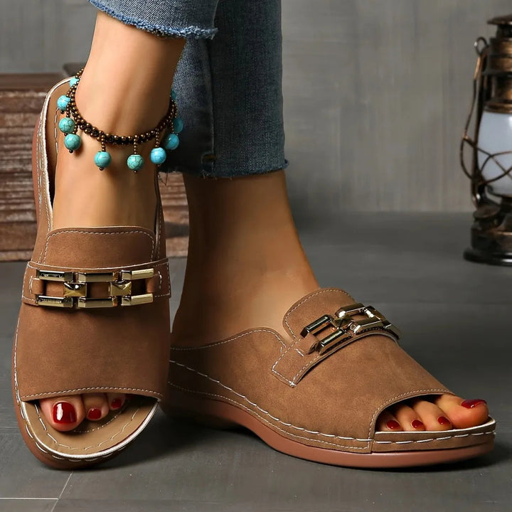Brown elegant sandals with gold details on feet, paired with blue anklet, set against a grey indoor background.