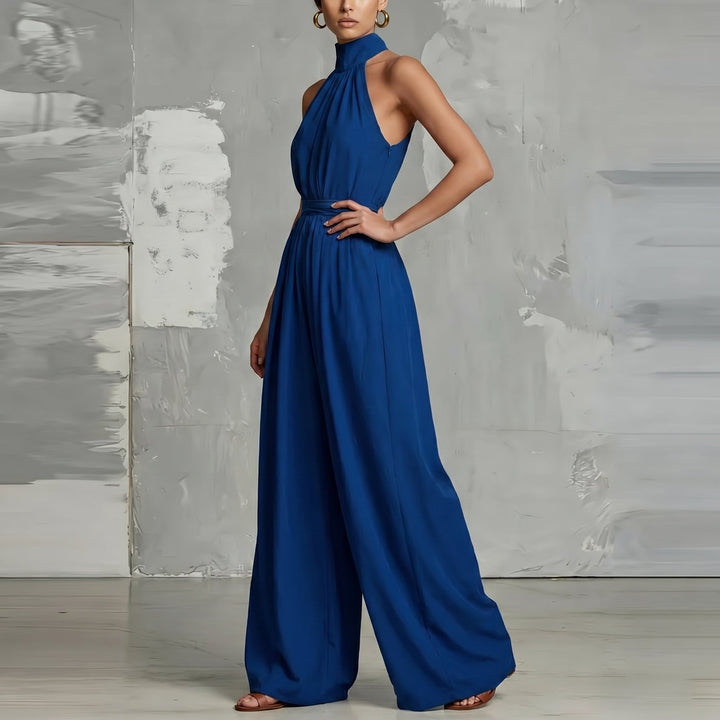 Blue halter-neck jumpsuit with wide legs, posed with hand on hip in a modern studio.