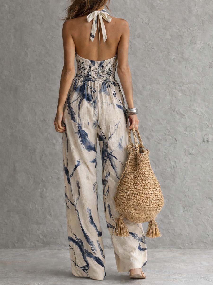 Back view of halter jumpsuit, smocked waist, blue print on beige, paired with boho bag. Textured studio backdrop.