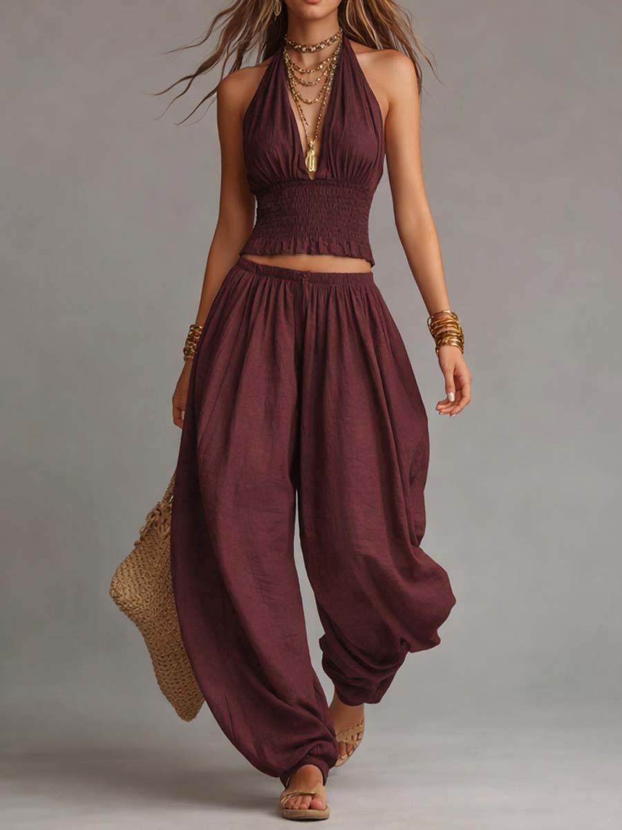 Maroon two-piece set with a halter top and airy pants, complemented by gold jewelry and a straw bag. Studio background.
