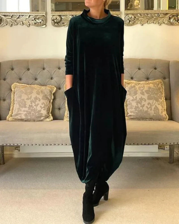 Dark green flowing suede dress with pockets, worn indoors. A tufted sofa with patterned pillows is in the background.