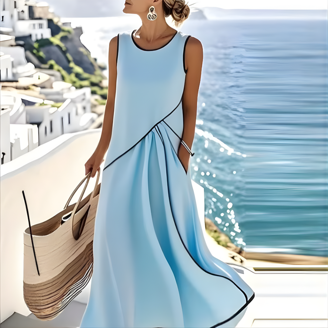 Woman in a flowing light blue summer dress with black trim, holding a large tote bag by the sea with white buildings in the background.
