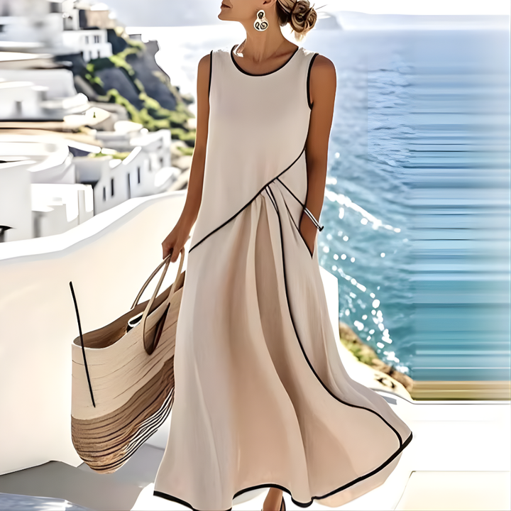 Woman in flowing cream summer dress with black trim, carrying a large tote bag against a seaside backdrop with white buildings.