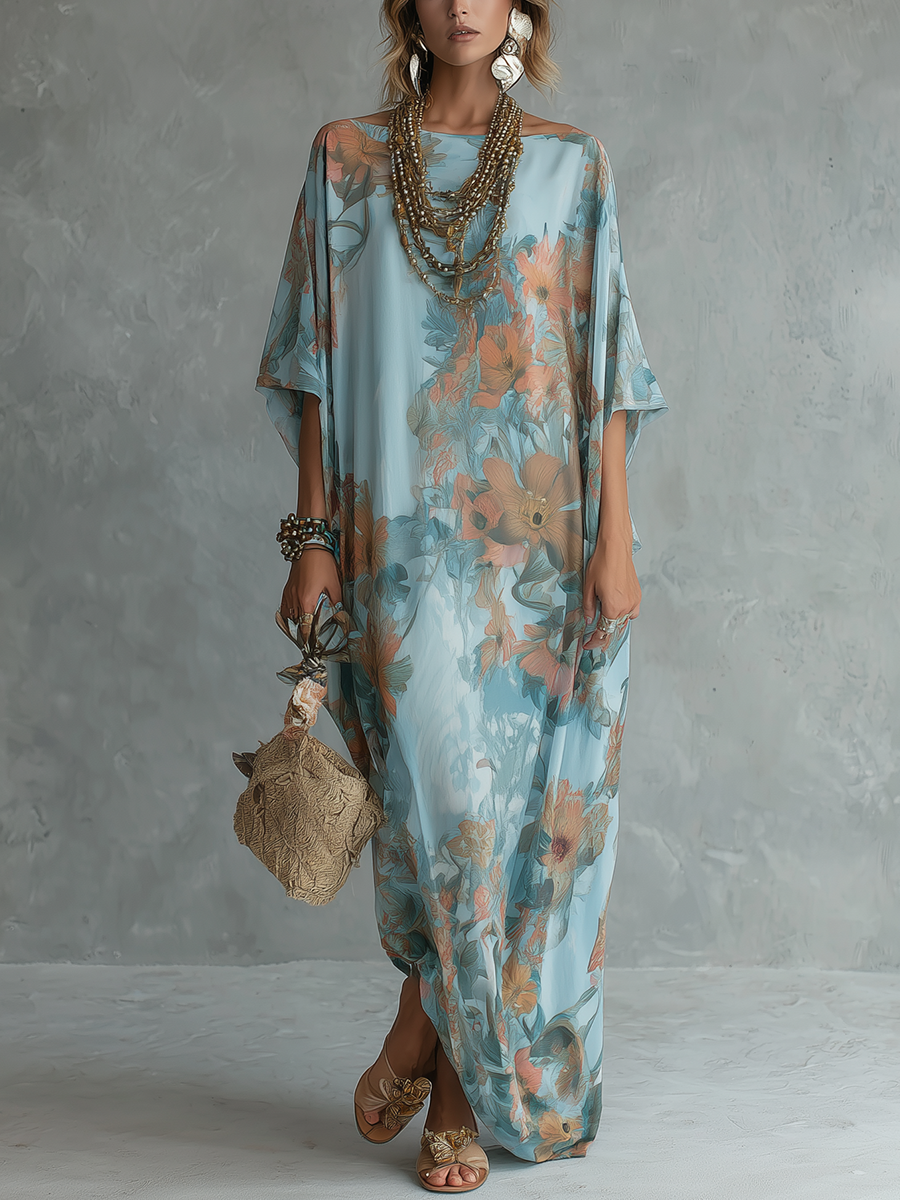 Model wearing a flowing floral long dress in shades of blue and orange, accessorized with layered necklaces, large earrings, a textured handbag, and sandals. Neutral textured studio backdrop.