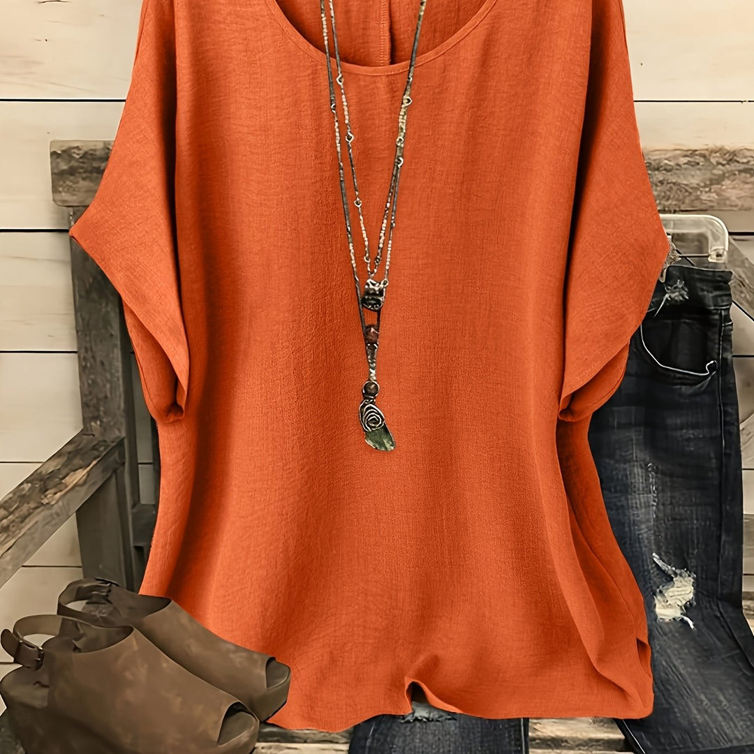Rusty orange loose summer top on hanger with necklace, sandals, and jeans on rustic bench.