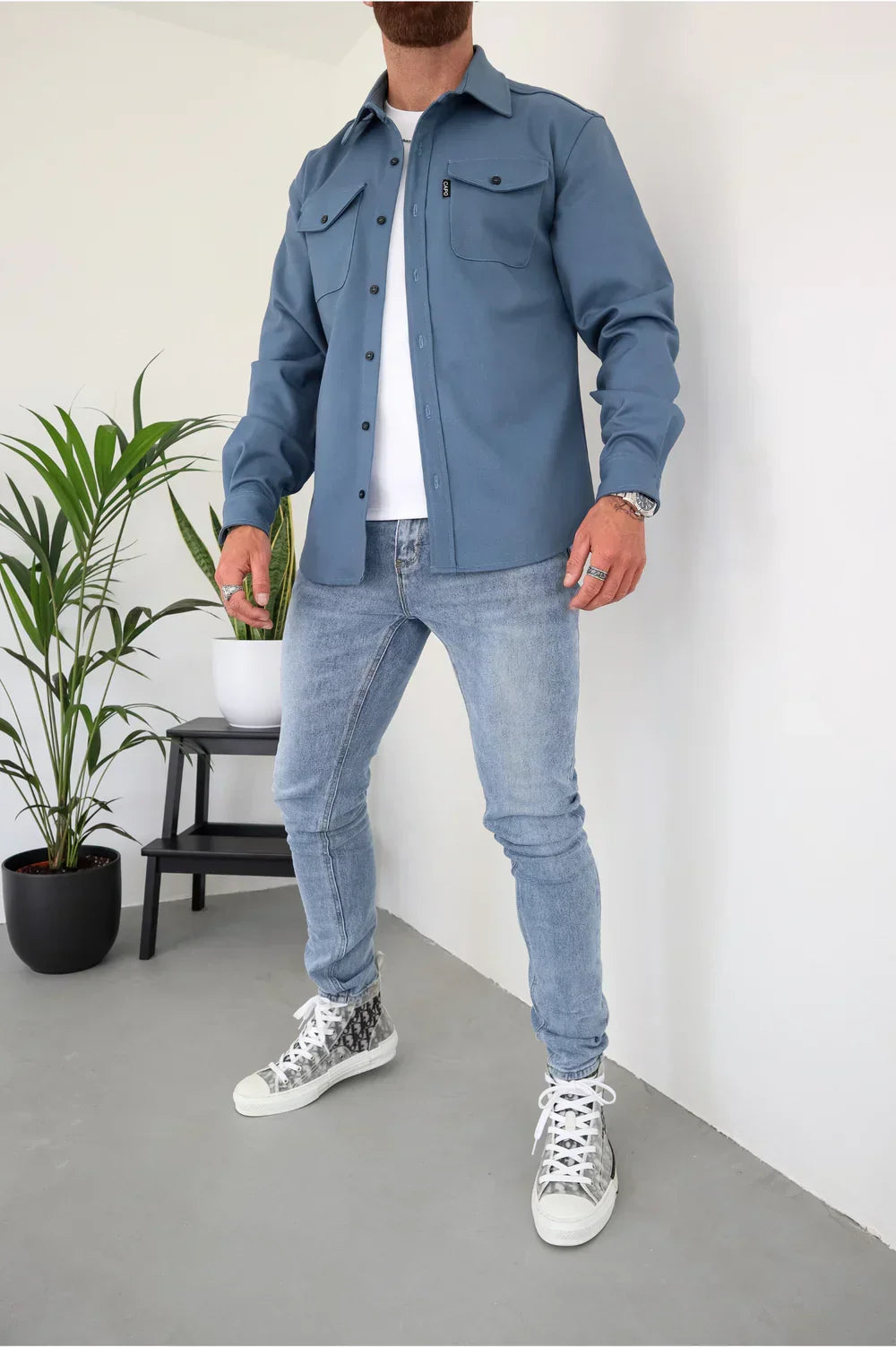 Blue overshirt over white t-shirt and light jeans, standing indoors beside plants and chair.