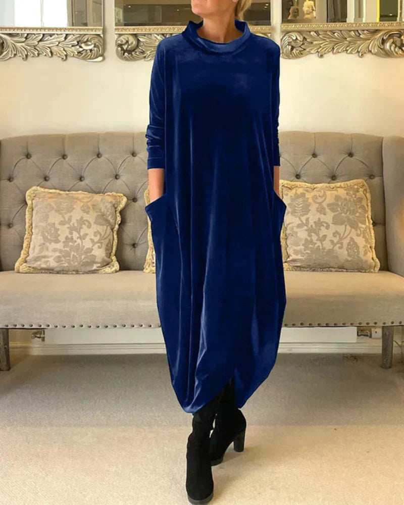 Blue flowing suede dress with pockets, showcased indoors with a tufted sofa and decorative pillows behind.