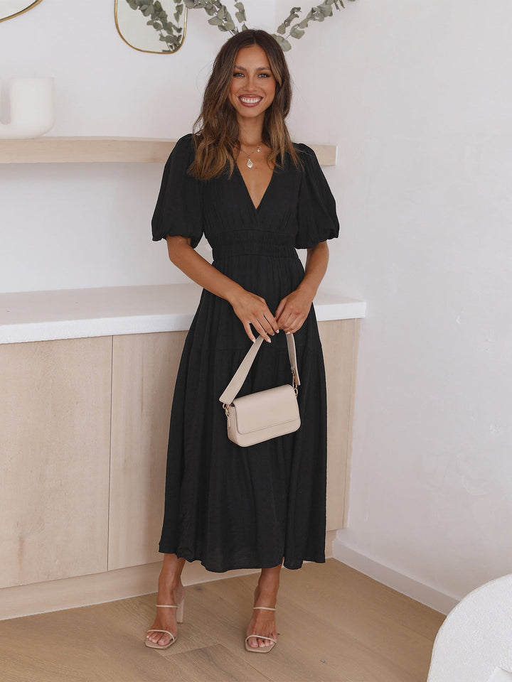 Woman in a black midi dress with puff sleeves, holding a beige bag in a stylish room.