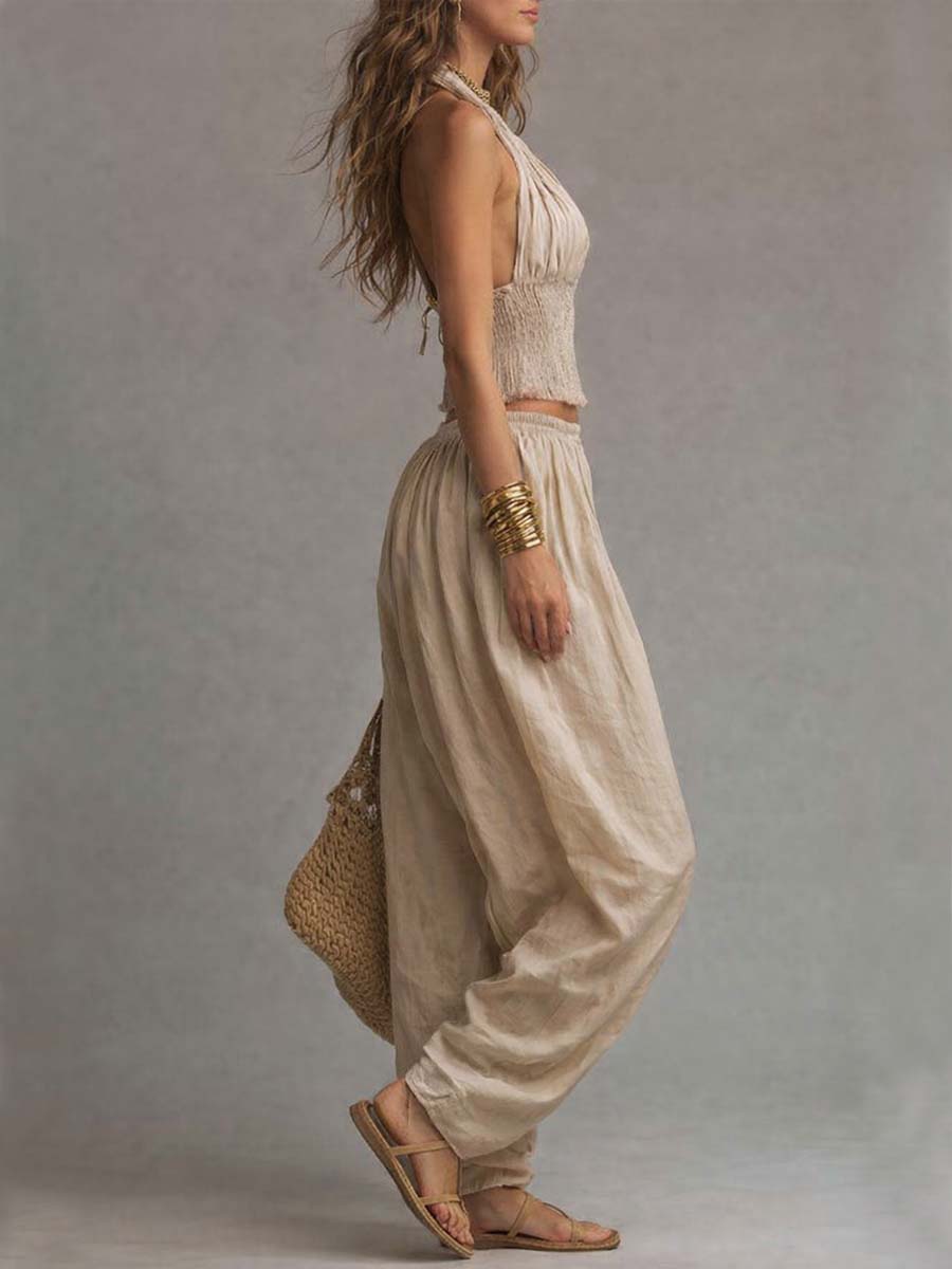 Side view of beige two-piece set featuring a halter top and relaxed pants. Woman carries a woven bag. Studio background.