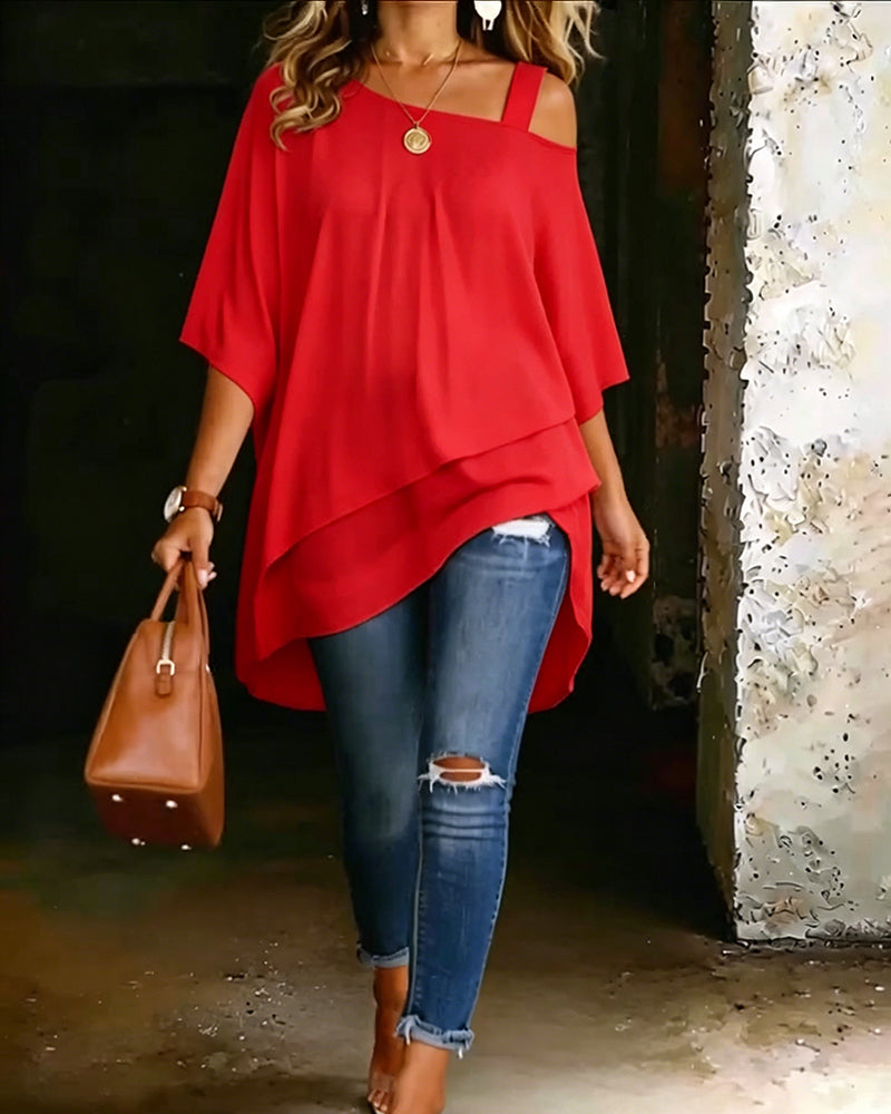Red asymmetrical blouse with off-shoulder straps paired with ripped jeans and brown handbag in rustic setting.