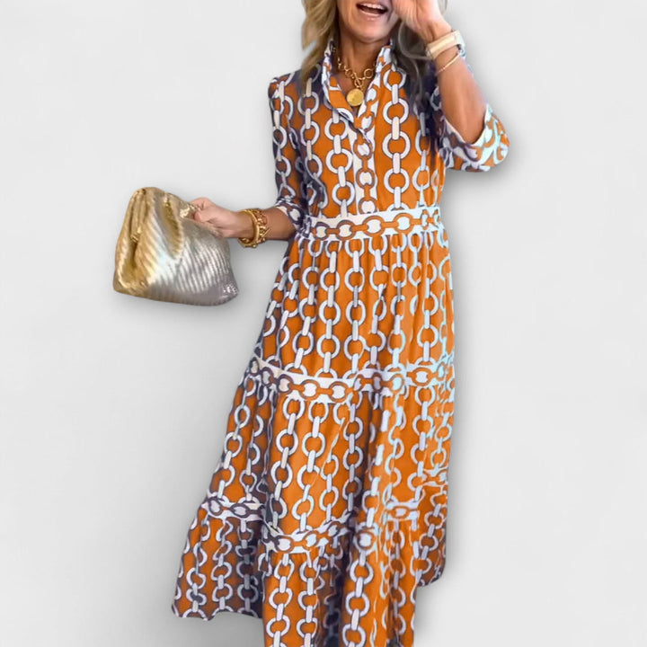 Woman in orange and white chain print midi dress holding a gold bag against a plain background.