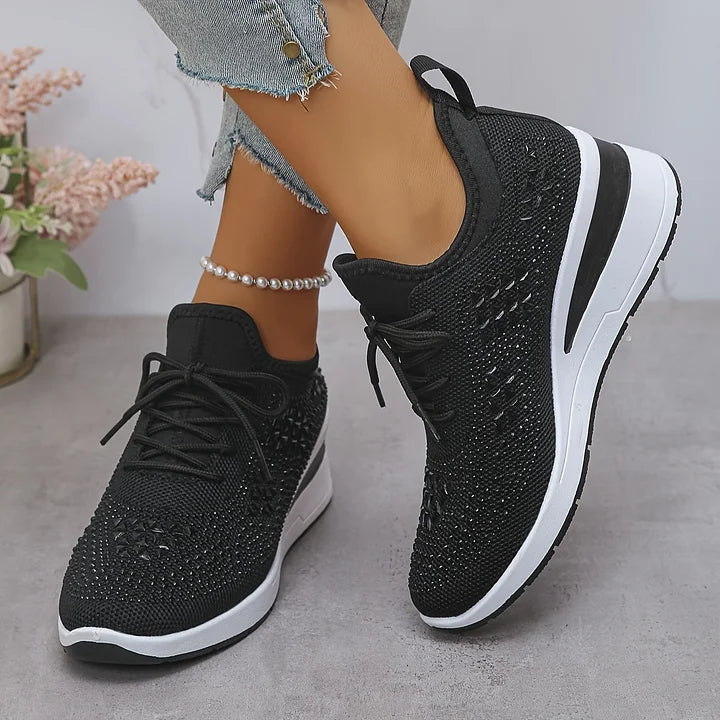 Black orthopedic sneakers with shimmering details, worn casually, set against a textured indoor surface.