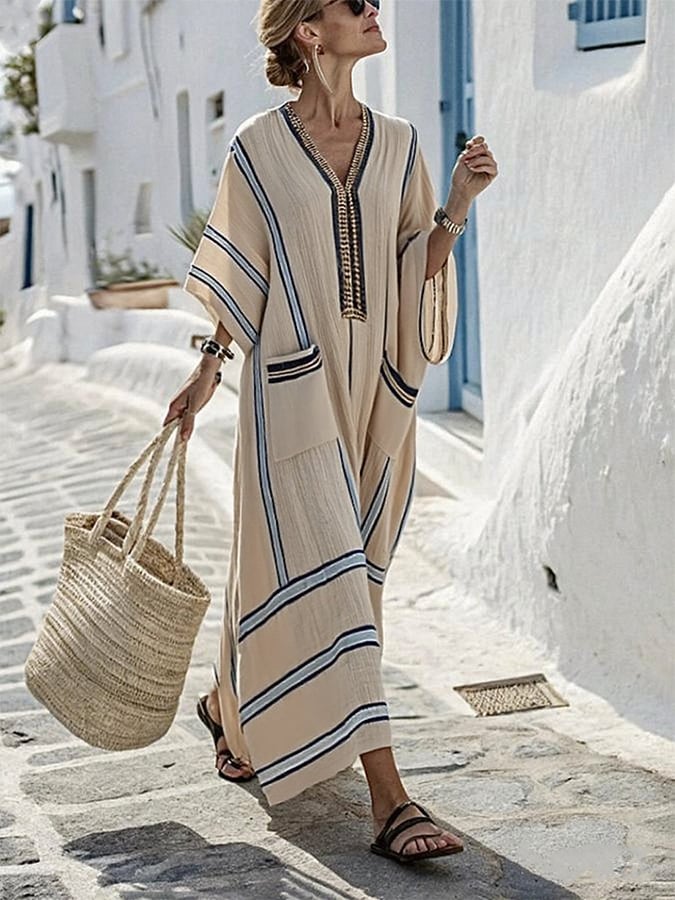 Elegant loose striped dress with pockets in beige and blue, worn by a woman on a sunny cobblestone street with a woven tote bag.