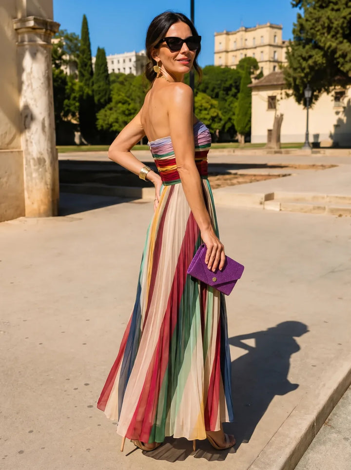 Back view of woman wearing a colorful pleated maxi dress, holding a purple clutch, standing outdoors in sunlight.