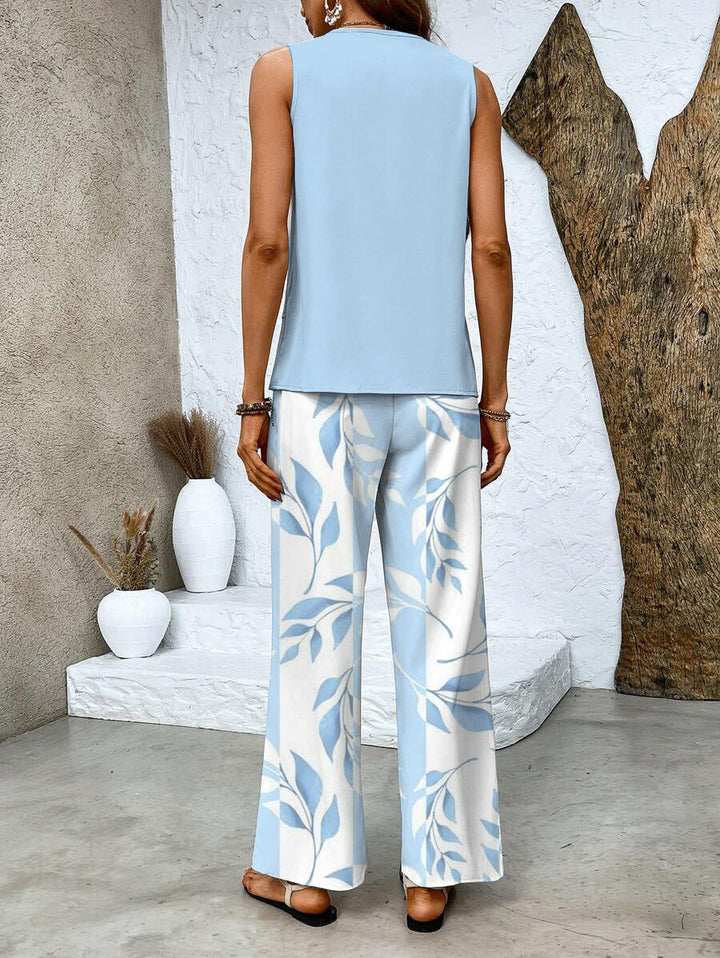 Back view of woman in light blue top and leaf print pants, standing near textured wall and white stairs with decorative vases.