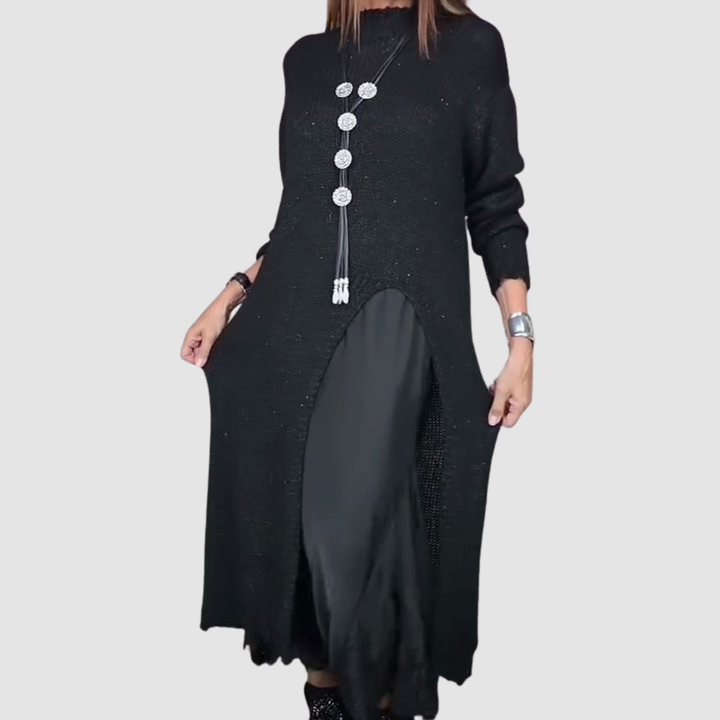 Woman in a black dress holding skirt sides, featuring a layered design and long necklace, on gray backdrop.