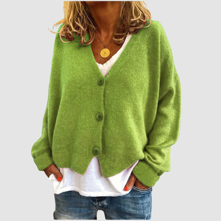 Woman wearing a light green knit cardigan over a white shirt and blue jeans, set against a plain gray background.