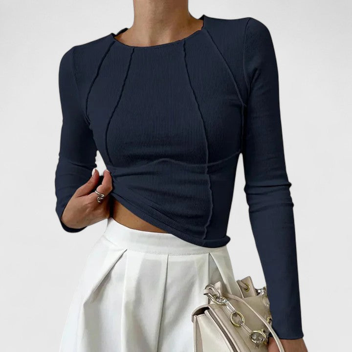 Dark blue long-sleeve top with curved seam details, worn with a white skirt and beige handbag, on a plain white background.