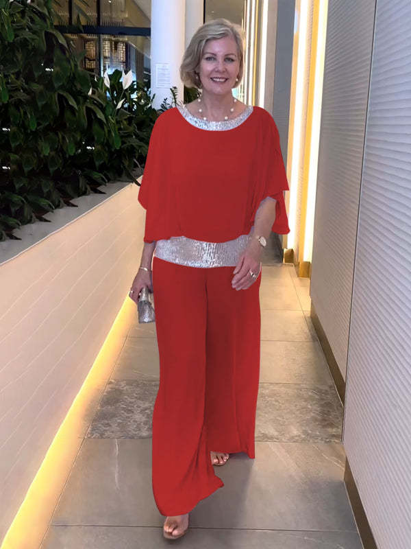 Woman in a red elegant set with shimmering belt and neckline, holding a silver clutch. Indoor setting with plants.