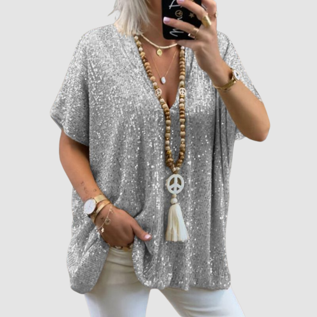 Woman wearing a silver V-neck sequin top with white pants, accessorized with layered necklaces and bracelets, on a neutral background.