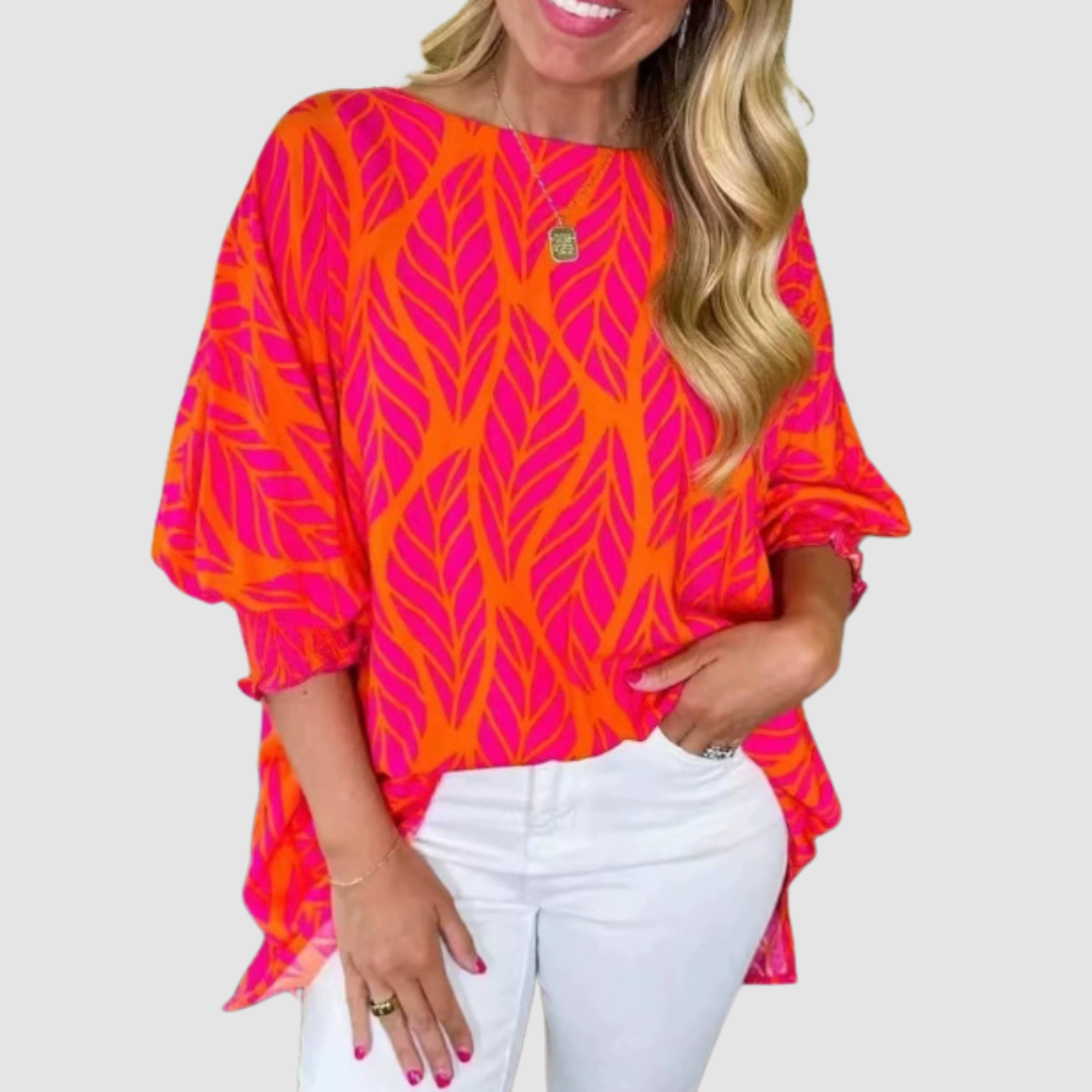 Woman wearing an orange and pink loose blouse with leaf pattern and white pants in a studio setting.