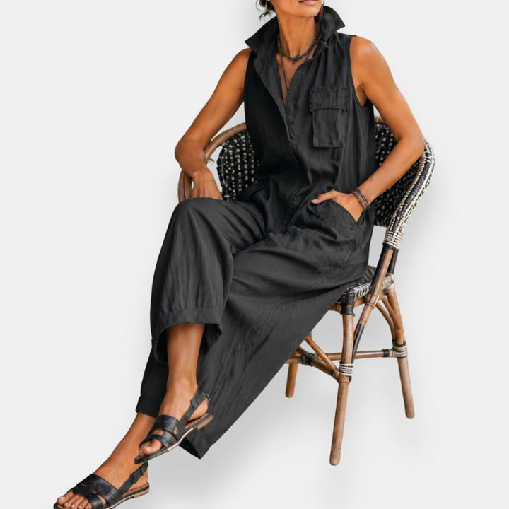 Model seated in black sleeveless cargo jumpsuit with collar and pockets on rattan chair against white background.