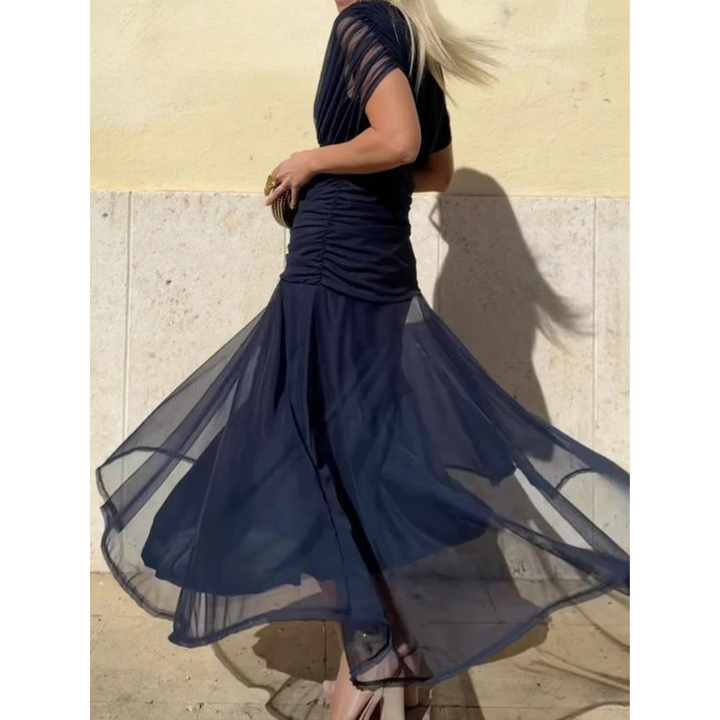 Woman in a navy blue mesh dress, holding a small clutch, standing against a light wall with shadows cast.