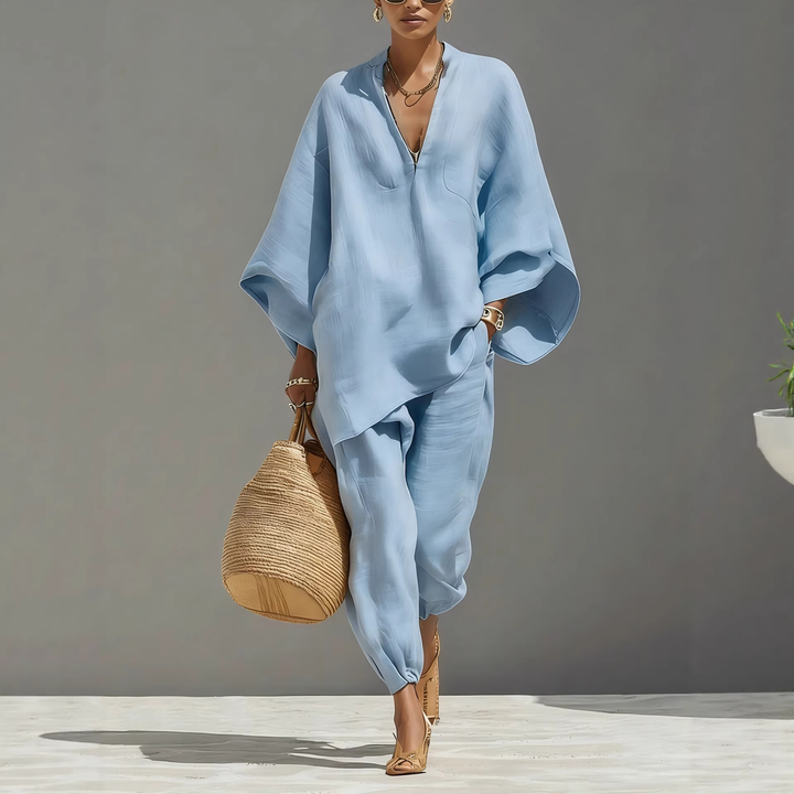 Model in light blue batwing sleeve V-neck lounge set holding straw bag, studio setting with plant.