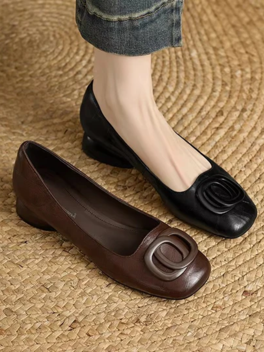 Black and brown leather flats with large buckles, worn with denim, on woven mat.