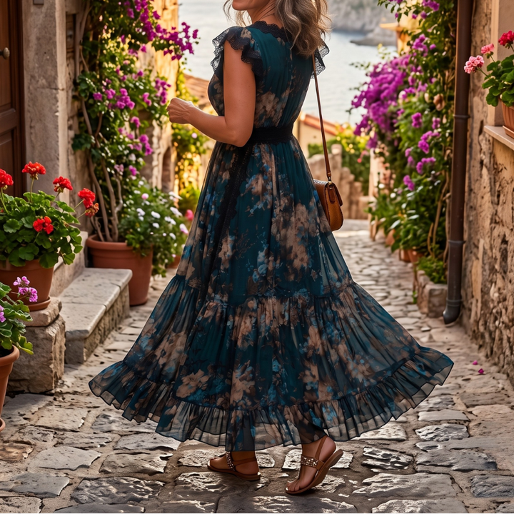 Back view of a woman in a flowing dark-patterned maxi dress on a cobblestone path with colorful flowers and a glimpse of the sea.