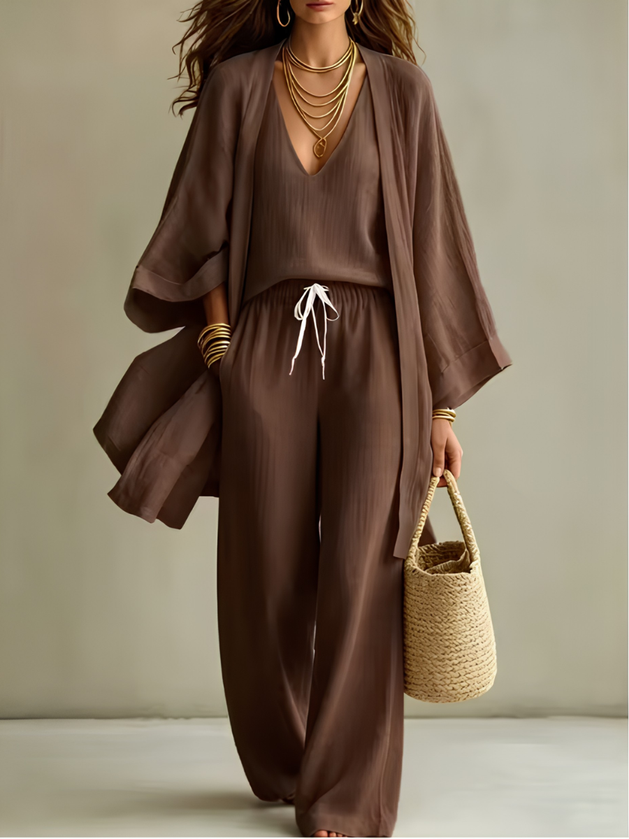 Brown three-piece relaxed homewear set with v-neck top, drawstring pants, flowy overcoat, and woven bag in studio setting.