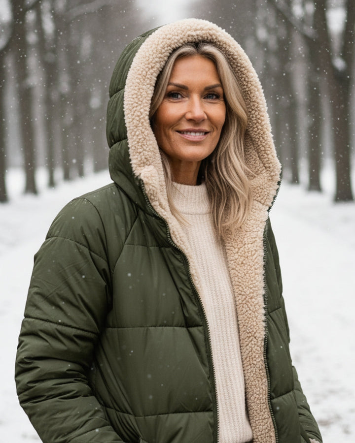 Woman in green hooded coat, smiling amidst snow on a tree-lined path.