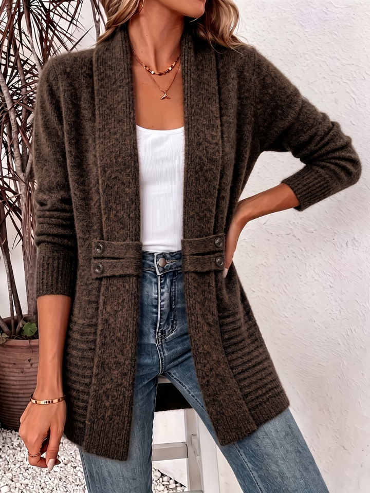 Woman in a dark brown knitted cardigan featuring buttons, with a white top and jeans, indoors against a white wall with decorative plants.