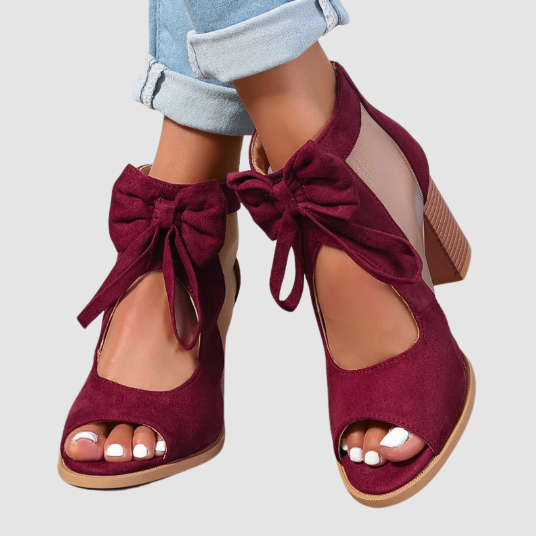 Burgundy heeled sandals with bow detailing, worn with cuffed jeans on a neutral background.