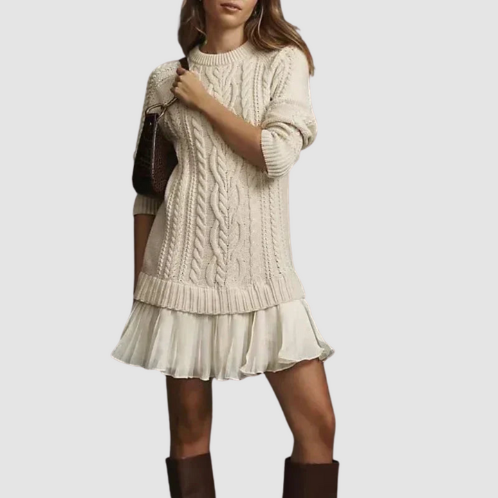 Cream cable knit mini dress with ribbed cuffs and a pleated hem, paired with a brown bag and boots. Studio backdrop.