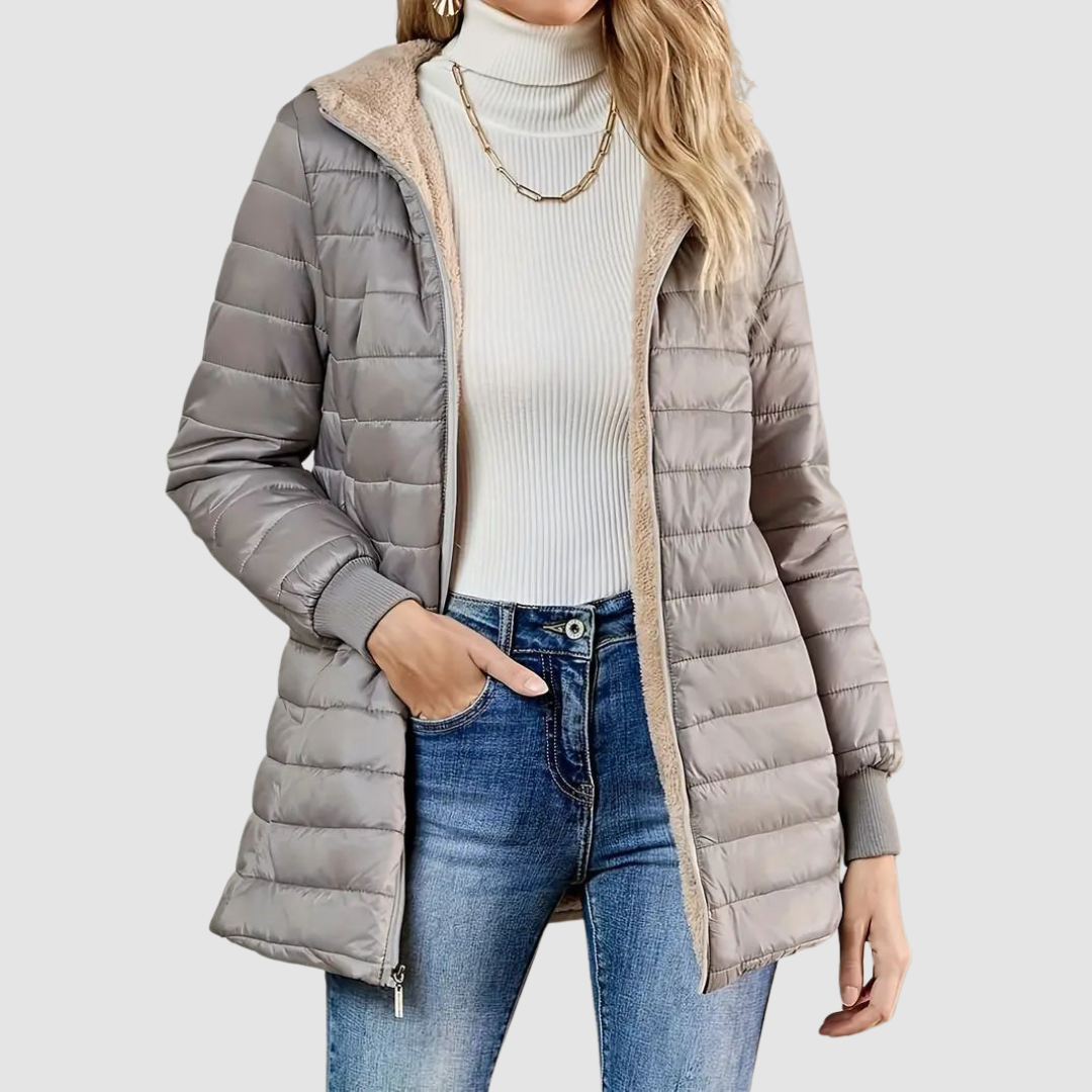 Woman wearing a light gray winter coat with a ribbed turtleneck, jeans, and a chain necklace, studio setting.