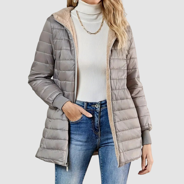 Woman wearing a light gray winter coat with a ribbed turtleneck, jeans, and a chain necklace, studio setting.