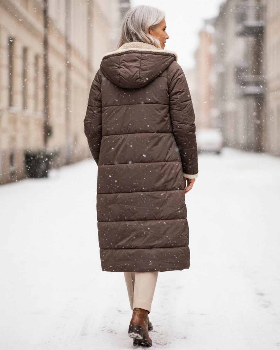 Back view of woman in long brown coat, walking down a snowy urban street.