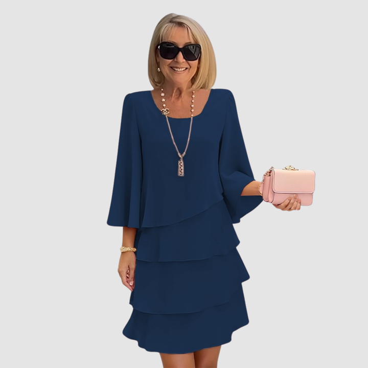 Elegant woman in a navy tiered dress with bell sleeves, holding a pink purse, on a light gray background.