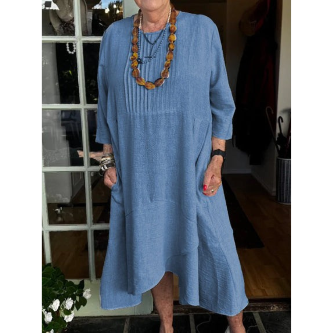 Woman in a blue pleated dress with irregular hem, wearing layered necklaces in a boutique setting.