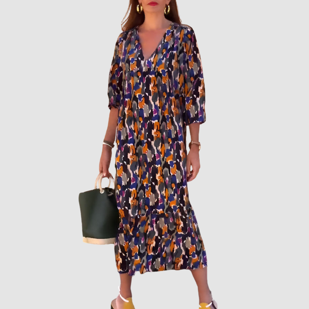 Model in a vibrant V-neck dress with abstract patterns, carrying a dark green bag, on a plain background.